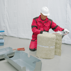 A worker in a red and black uniform with a white hard hat kneels on a floor while handling large rolls of beige fibrous insulation material, with metal trim pieces and tools nearby against a white backdrop.