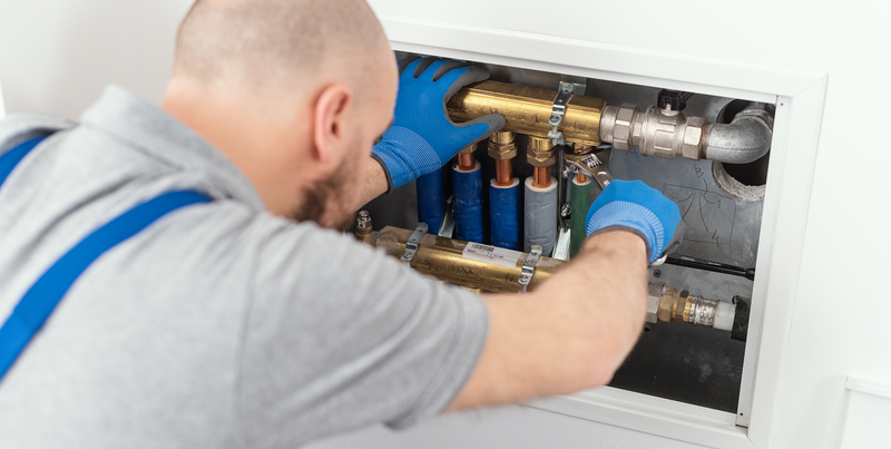 A plumber in a gray shirt with blue suspenders and blue gloves works inside a white wall-mounted utility cabinet, adjusting brass and copper pipes with metal clamps.